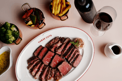 Three cuts of steak grilled on open wood fire grill with bottle of wine and some sides